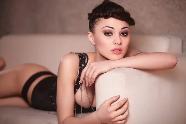 Menina de moda sexy bonita em espartilho preto deitado no sofá dentro de casa maquiagem brilhante e penteado — Fotografia de Stock