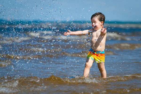 Ein süßer kleiner Junge, der am Strand durchs Wasser rennt — Stockfoto