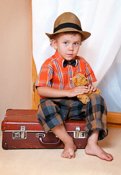 Ένα αγόρι με ένα αρκουδάκι που κάθεται σε μια βαλίτσα. — Φωτογραφία Αρχείου