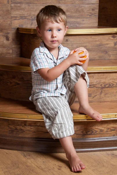 Um menino bonito com uma laranja na mão sentado nos degraus . — Fotografia de Stock
