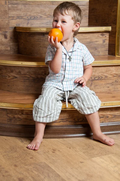Um menino bonito com uma laranja na mão sentado nos degraus . — Fotografia de Stock