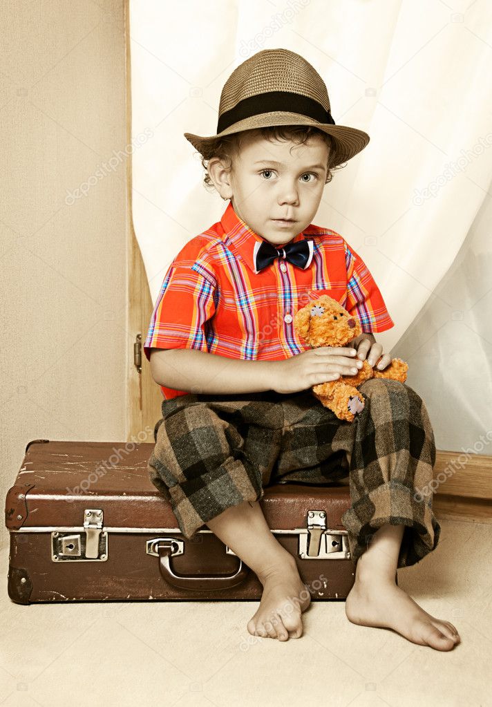 A boy with a teddy bear sitting on a suitcase.
