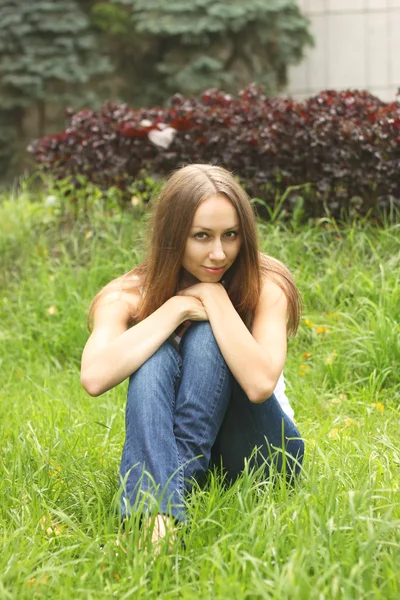 La muchacha en los vaqueros azules oscuros se sienta sobre la hierba — Foto de Stock