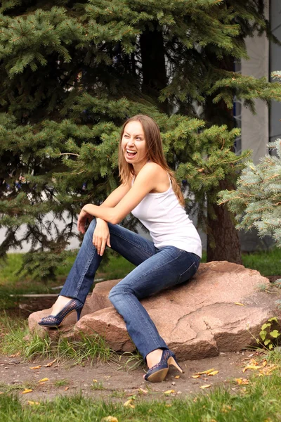 Das Mädchen in weißer Weste und dunkelblauer Jeans sitzt auf Steinen im Quadrat — Stockfoto