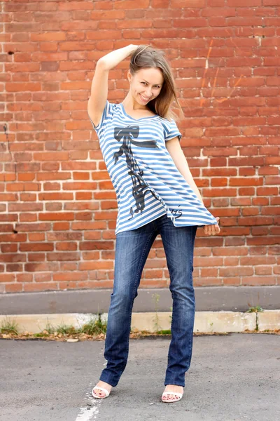 The girl in a striped vest and dark blue jeans costs at a brick wall — Stock Photo, Image