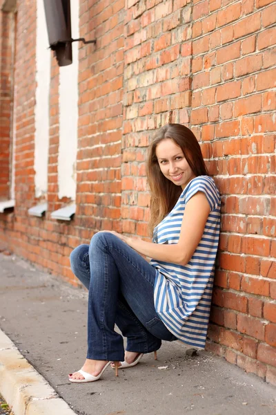 Dívka v pruhované vestě a tmavě modré džíny sedí na cihlovou zeď — Stock fotografie