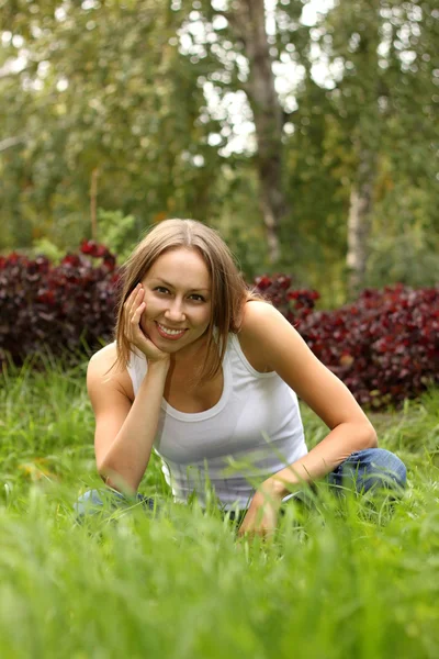 Het mooie meisje in een wit vest en blue jeans zit op een gras — Stockfoto
