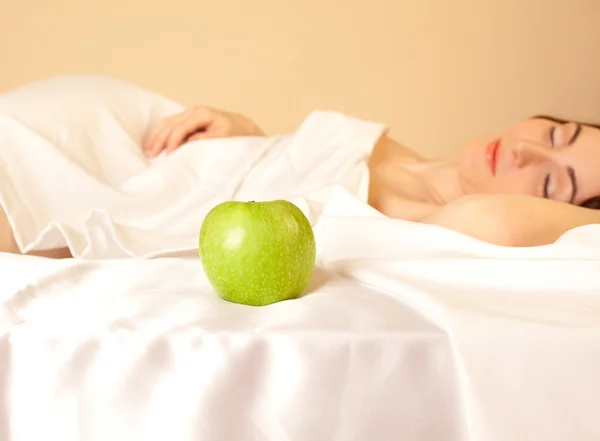 Beautiful woman lying in bed with apple (focus on apple) — Stock Photo, Image