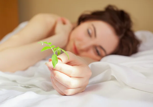 Beautiful woman holding a plant and lying in bed (focus on sprou — Stock Photo, Image