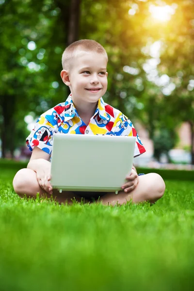 Mignon petit garçon assis avec un ordinateur portable sur l'herbe — Photo