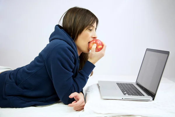 Женщина ест красное яблоко перед компьютером лежа в постели — стоковое фото