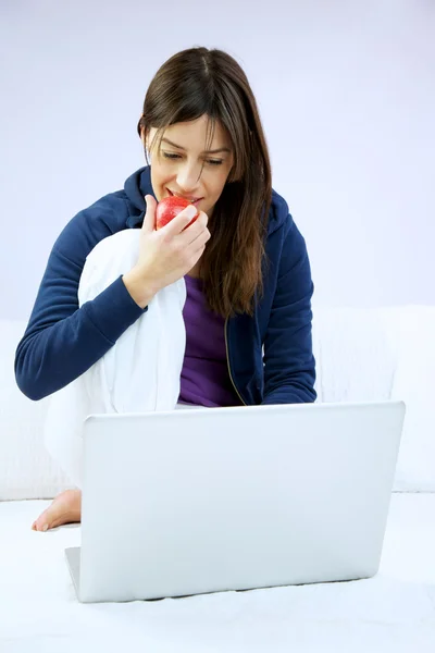 Woman smiling eats apple sitting in front of computer — Stock Photo, Image