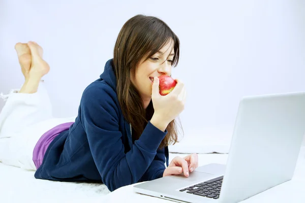 Улыбающаяся женщина ест яблоко перед компьютером, лежащим в постели — стоковое фото
