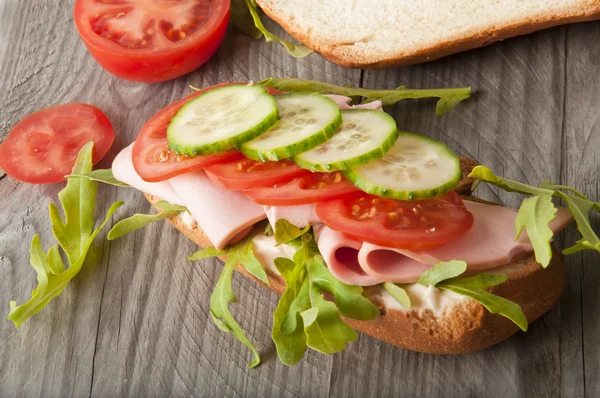 Sandwich met ham, tomaat, komkommer en rucola op de oude houten snijplank — Stockfoto