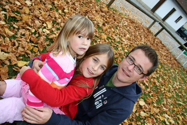 Father with his two daughters sitting in a park in autum leaves — Stock Photo, Image