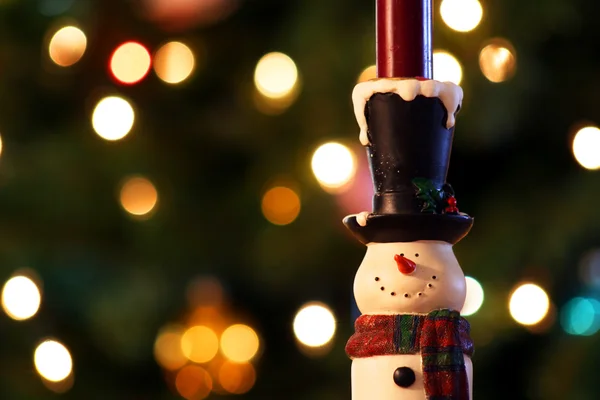 A cheerful snowman candle holder stands before the colored lights of a Chri — Stock Photo, Image