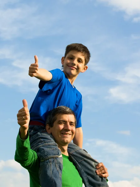 Un niño está sentado en el cuello de su padre sosteniendo sus pulgares hacia arriba — Foto de Stock