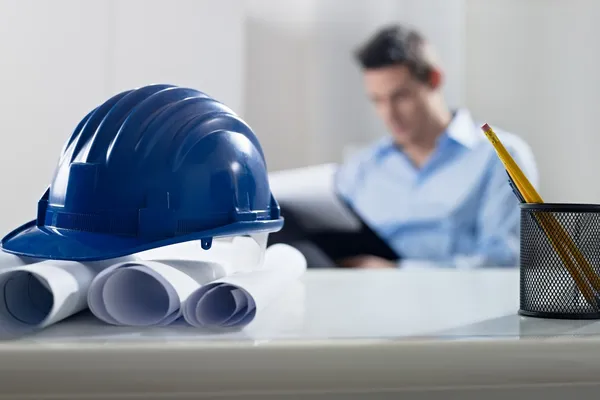Hardhat und Blaupause auf dem Schreibtisch, mit Architekt im Hintergrund — Stockfoto