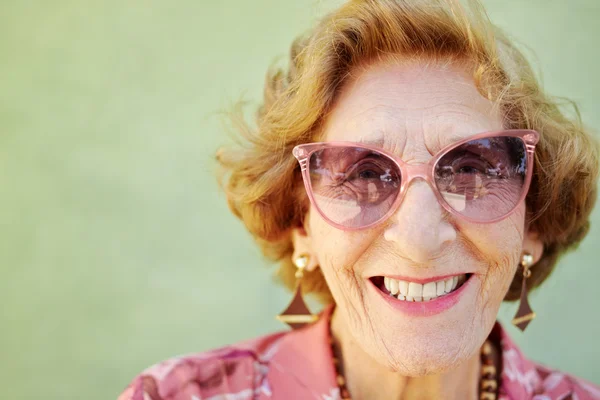 Pembe gözlükler kameraya gülümseyen yaşlı kadınla — Stok fotoğraf