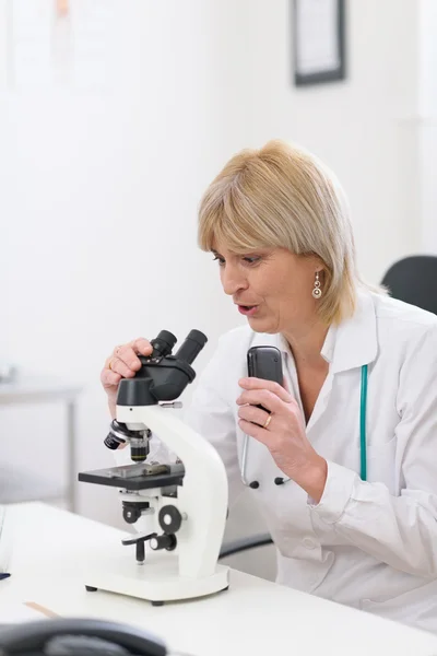 Mujer doctora de mediana edad sorprendida mirando en el microscopio y haciendo notas de voz — Foto de Stock