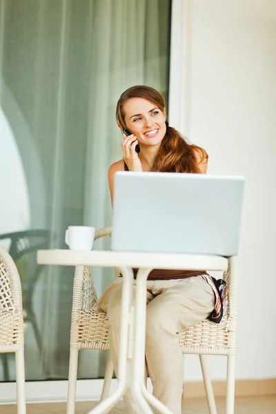 Donna sorridente sulla terrazza che parla cellulare e lavora sul computer portatile — Foto Stock