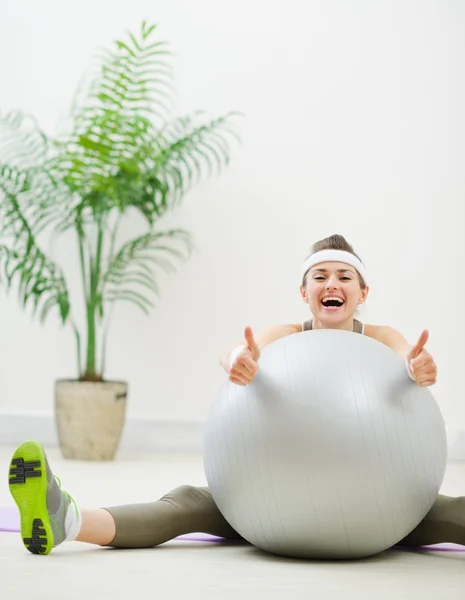 Smiling woman sitting behind fitness ball and showing thumbs up — Stock Photo, Image