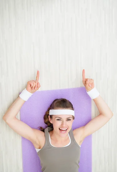 Mujer joven feliz acostada en la alfombra de fitness y apuntando en el spa de copia —  Fotos de Stock
