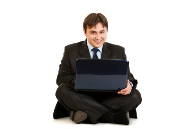 Smiling businessman sitting on floor and working on laptop clipart