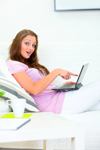 Mujer bonita confundida sentada en el sofá y señalando con el dedo a la computadora portátil — Foto de Stock