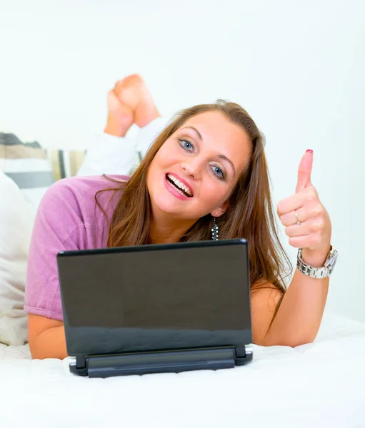 Sorrindo mulher bonita deitada no sofá com laptop e mostrando polegares para cima gestur — Fotografia de Stock
