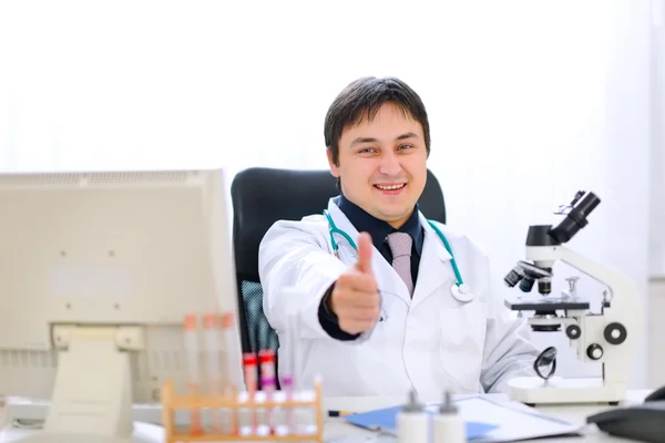 Médico sonriente sentado en la mesa del consultorio y mostrando los pulgares hacia arriba ges —  Fotos de Stock