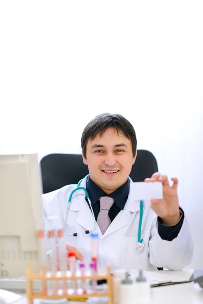 Médico sonriente sentado en la mesa del consultorio y sosteniendo negocios en blanco — Foto de Stock