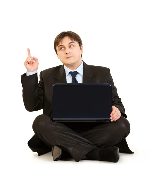 Задумчивый бизнесмен сидит на полу с ноутбуком и показывает пальцем вверх . — стоковое фото