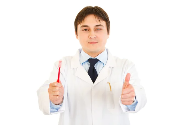 Dentista sonriente sosteniendo el cepillo de dientes y mostrando los pulgares hacia arriba gesto —  Fotos de Stock