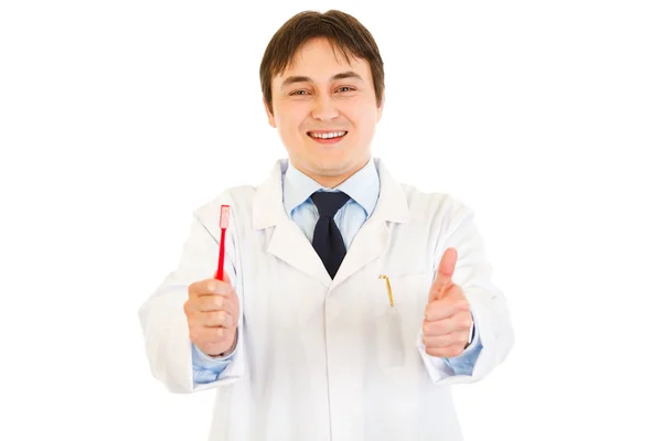 Dentista sonriente sosteniendo el cepillo de dientes y mostrando los pulgares hacia arriba gesto —  Fotos de Stock