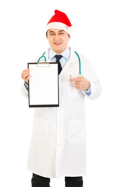 Uśmiechający się lekarza w santa hat wskazując palcem na puste schowka — Zdjęcie stockowe