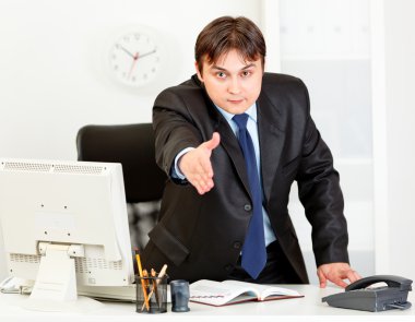 Friendly modern businessman standing at office desk and stretches out hand clipart