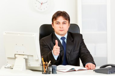 Confident modern businessman sitting at office desk and showing thumbs up g clipart