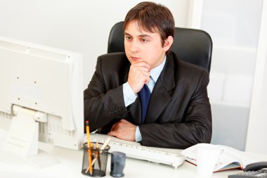 Concentrated businessman sitting at office desk and looking at computer mon clipart