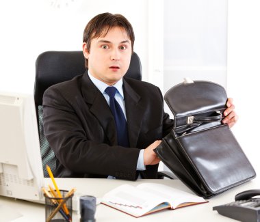 Shocked modern businessman sitting at office desk and holding open suitcase clipart