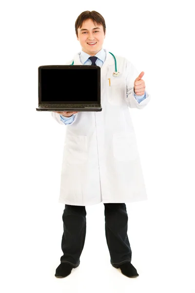 หมอที่ยิ้มถือแล็ปท็อปที่มีหน้าจอว่างและแสดงนิ้วโป้งขึ้น Ge — ภาพถ่ายสต็อก