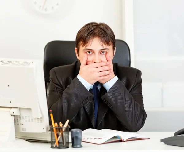 Modern businessman sitting at office desk and making speak no evil gesture — Stock Photo, Image