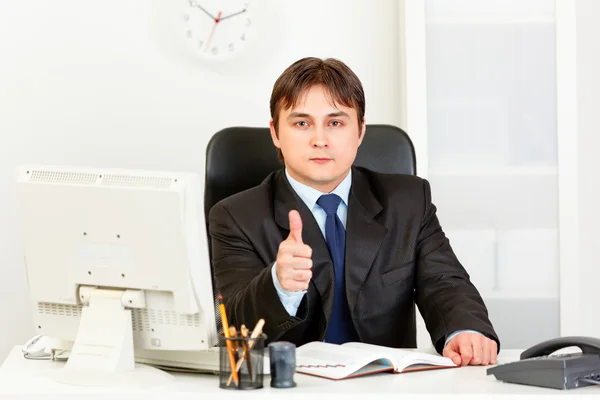 Confiante homem de negócios moderno sentado na mesa do escritório e mostrando polegares para cima g — Fotografia de Stock