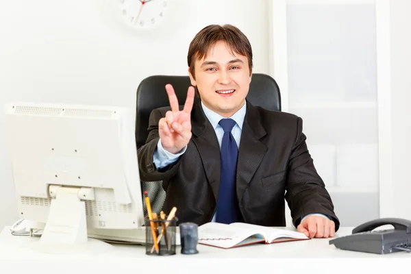 Sorridente uomo d'affari moderno seduto alla scrivania dell'ufficio e mostrando gestu vittoria — Foto Stock