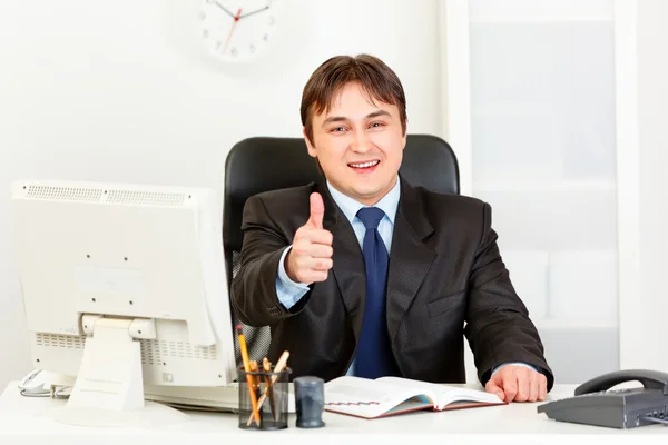Sorridente uomo d'affari moderno seduto alla scrivania dell'ufficio e mostrando pollici in su ges — Foto Stock