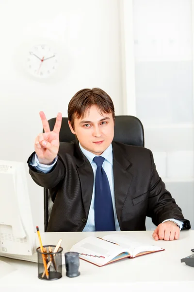 Confiado hombre de negocios moderno sentado en el escritorio de la oficina y mostrando ges victoria — Foto de Stock