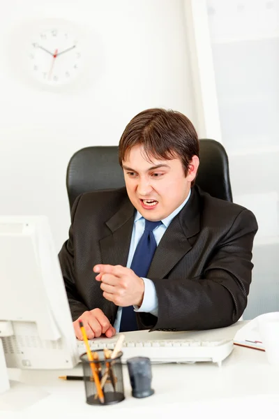 Angry businessman sitting at office desk and pointing finger at computer mo — Stock Photo, Image