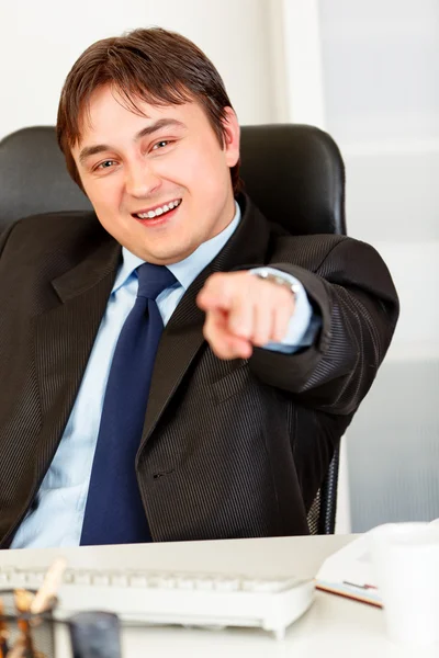 Empresário sorridente sentado na mesa do escritório e apontando o dedo para você — Fotografia de Stock