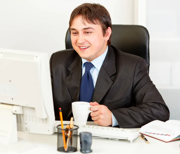 Uomo d'affari ridente con una tazza di caffè guardando il monitor del computer — Foto Stock
