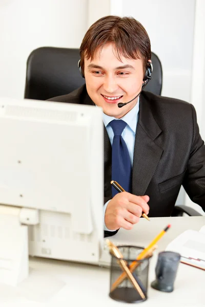 Empresário sorridente com fone de ouvido sentado na mesa do escritório e olhando para co — Fotografia de Stock